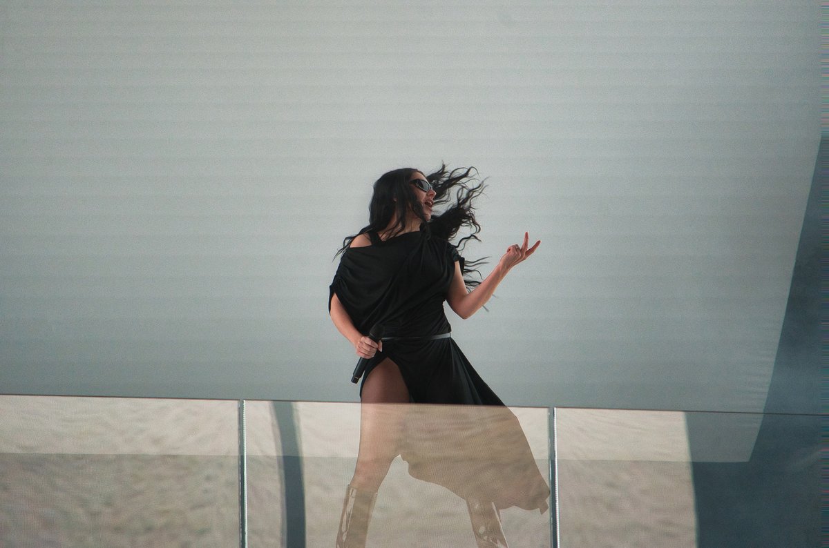 Charli XCX performing in an all-black outfit and sunglasses, mid-motion with her hair in the air, against a plain pale grey wall.