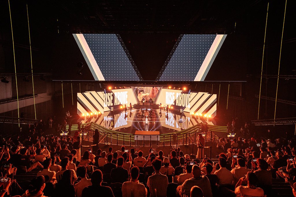 A packed Esports World Cup arena, phones raised, in front of a giant stage reading Rise Above